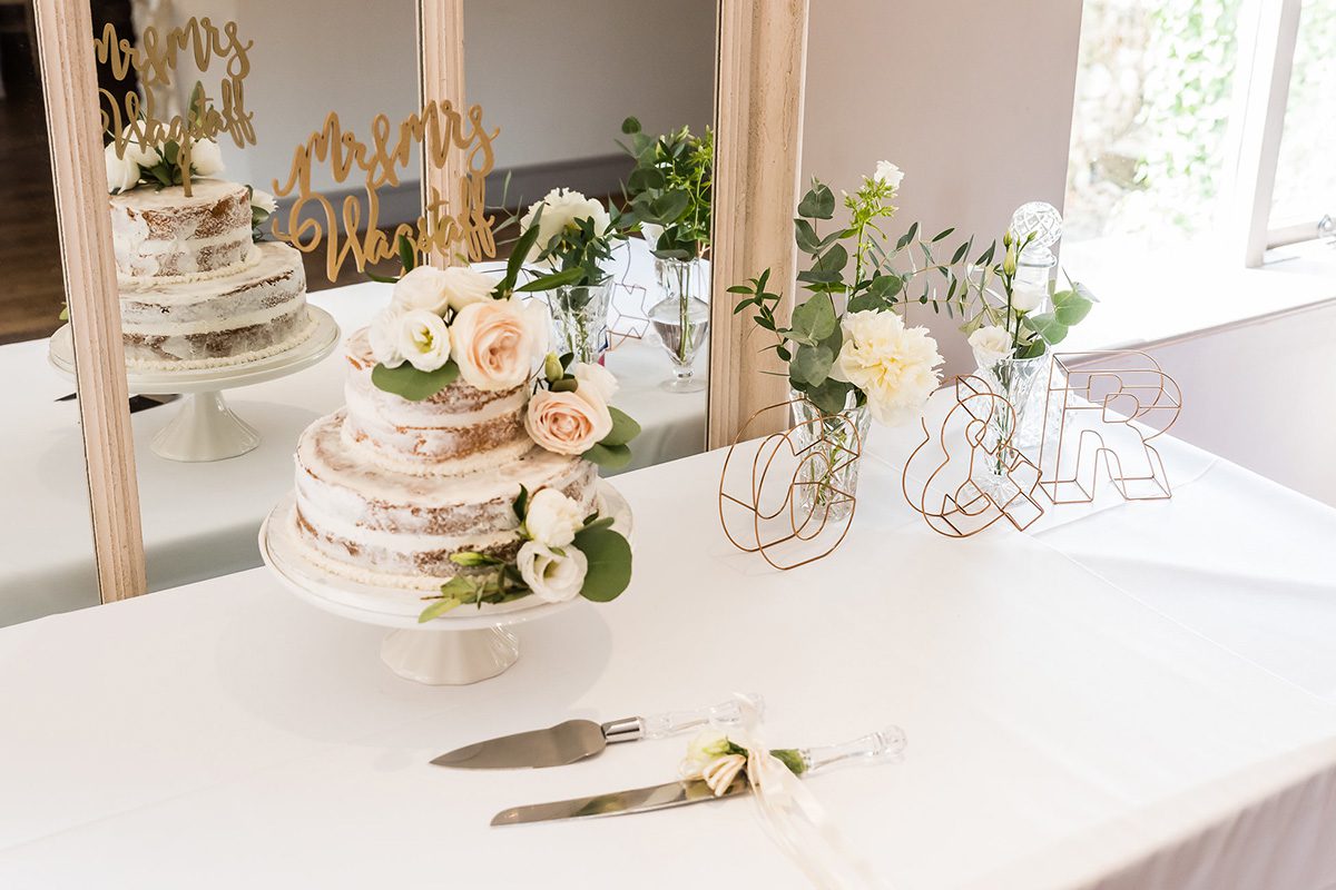 A beautiful three-tier semi-naked wedding cake decorated with pretty pale roses and a rose gold cake topper at Pentney Abbey